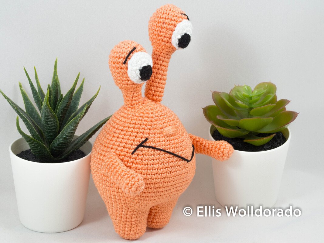 Orangenes gehäkeltes Amigurumi mit zwei Augenstielen zwischen zwei Sukkulenten