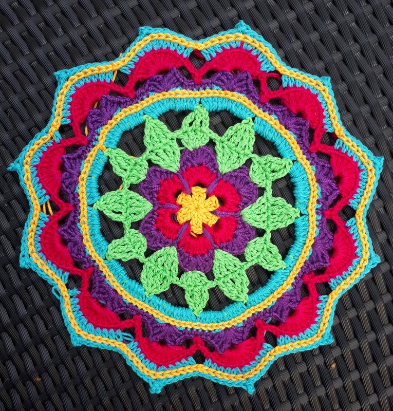 Runder gehäkelter Mandala-Untersetzer mit Blumen- und Blättermotiv in Pink, Lila, Grün, Türkis und Gelb auf dunklem Untergrund