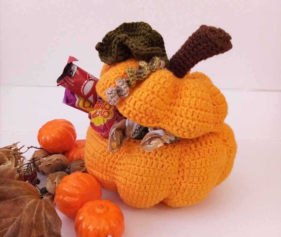 Hand-crocheted orange pumpkin container with removable lid, filled with wrapped candies, surrounded by small decorative pumpkins and dried leaves.
