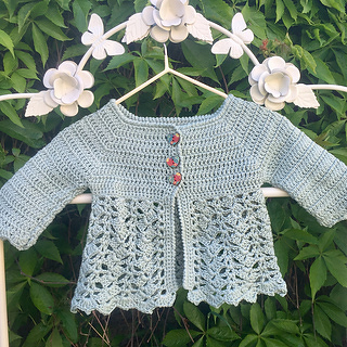 Pastel green crocheted baby cardigan with scalloped lace hem, textured yoke and three small buttons on a flower-adorned hanger