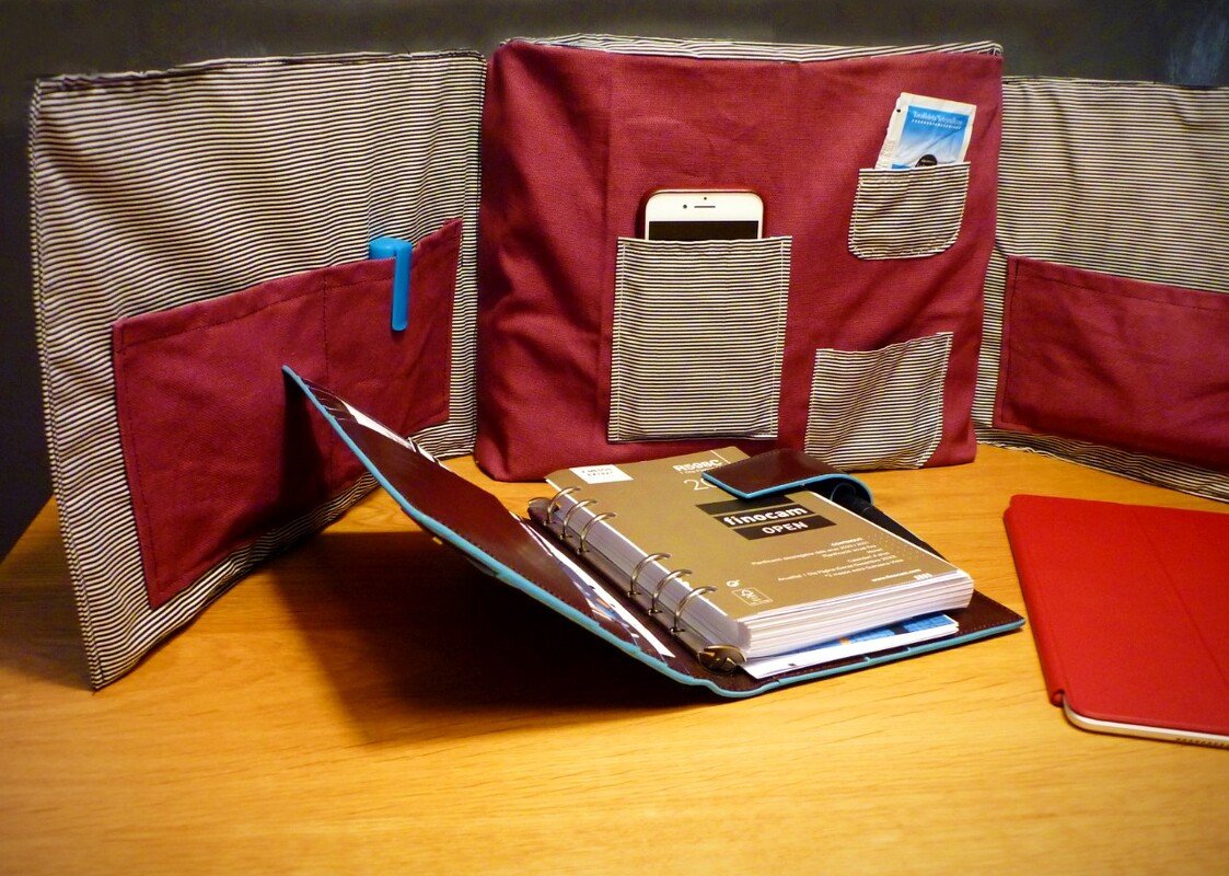 Drei bordeauxrote Stofforganizer mit gestreiften Taschen, bestückt mit Smartphone und Stift, neben einem Ringordner und Tablet auf Holz