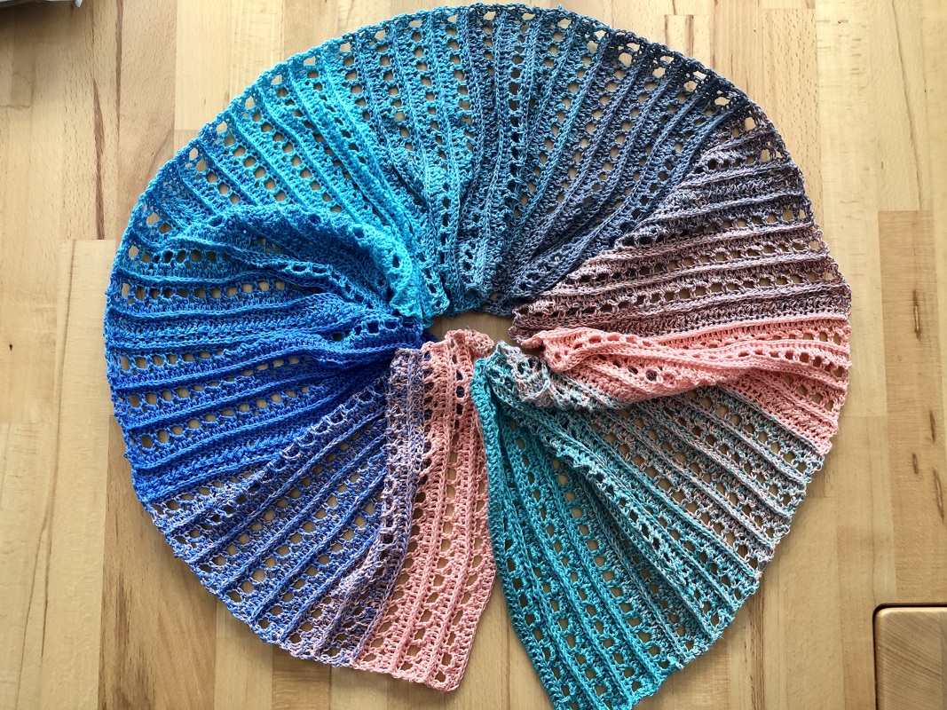 Halbrunder gehäkelter Schal mit Farbverlauf in Türkis, Blau, Grau und Rosa, aufgefächert auf hellem Holzboden