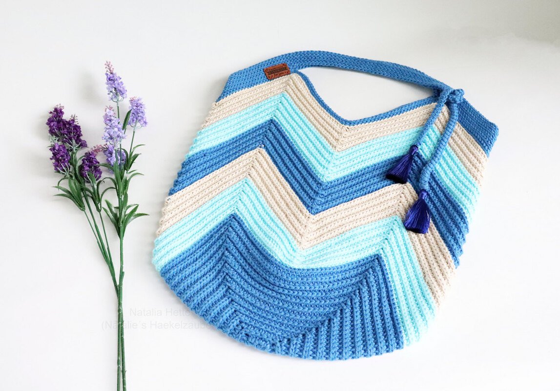 Chevron crochet handle bag in blue and beige with strap and tassels beside lavender on white.