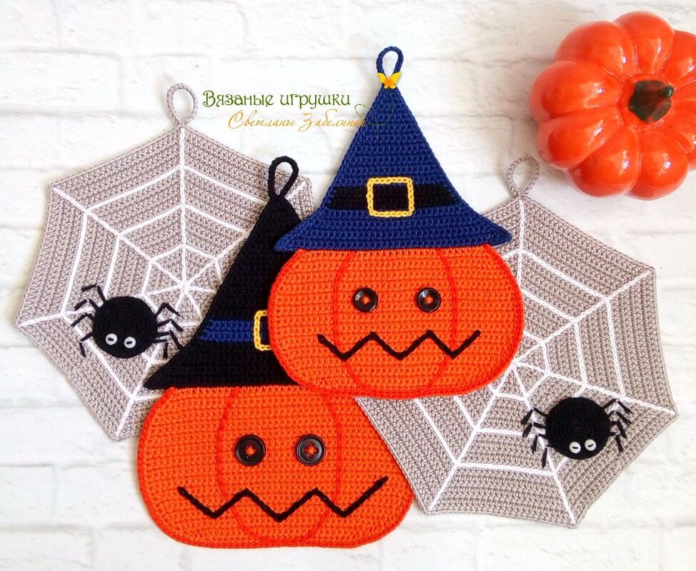 Gehäkelte orange Kürbisse mit blauen Hexenhüten neben hellgrauen Spinnennetz-Topflappen mit schwarzen Spinnen