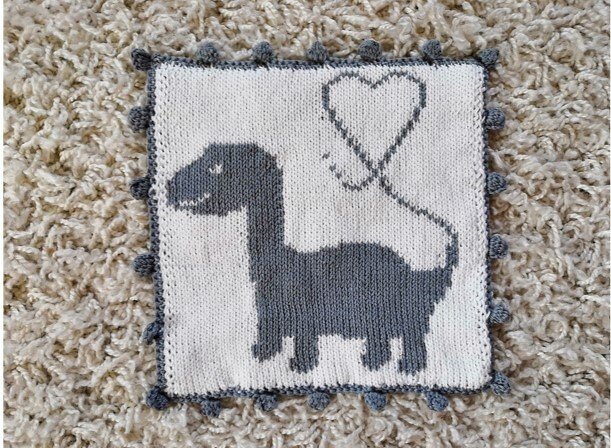 Gestricktes Quadrat in Weiß und Grau mit Dinosaurier- und Herzmotiv und Bommelrand