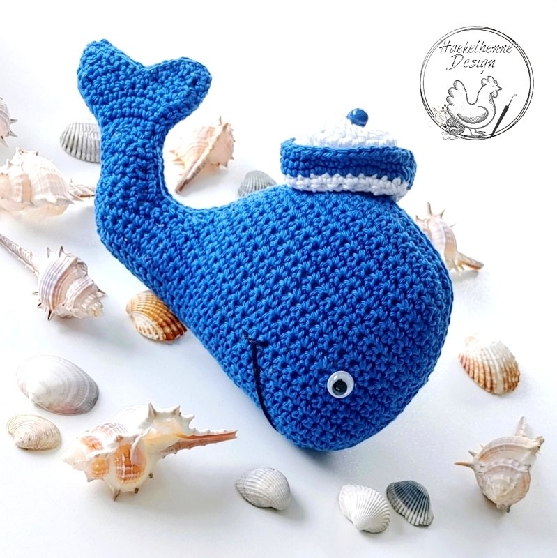 Blauer gehäkelter Amigurumi-Wal mit weiß-blauer Mütze und Wackelauge, umgeben von Muscheln