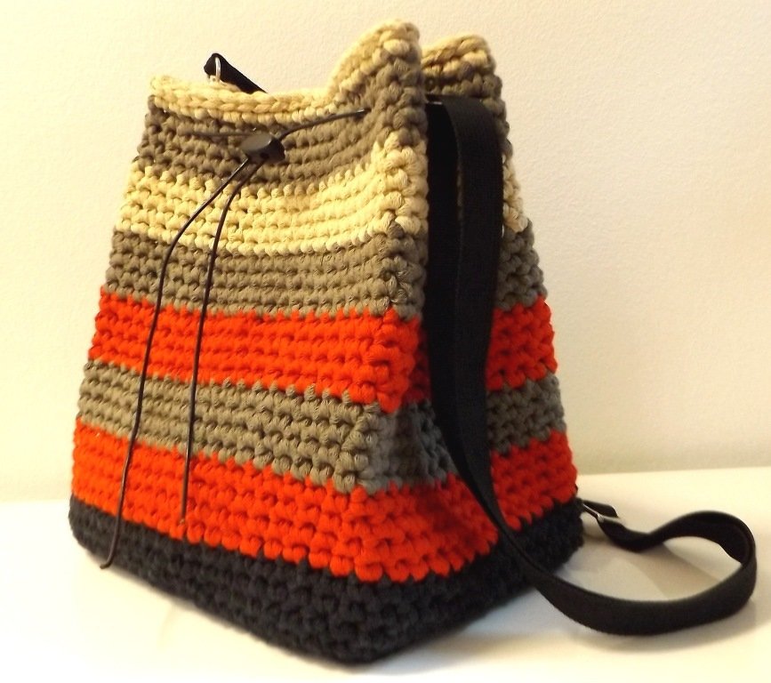 Gehäkelte Beuteltasche mit horizontalen Streifen in Beige, Grau und Orange, Kordelzug und dunklem Umhängegurt.