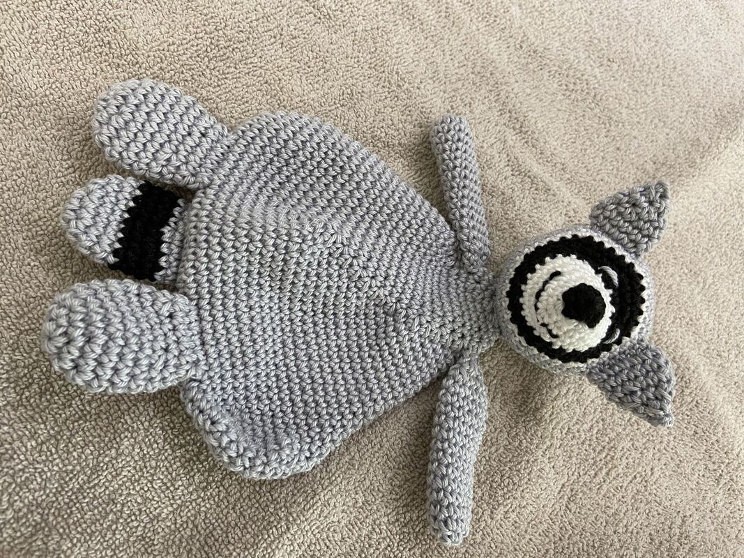 Gehäkeltes graues Schmusetuch mit Waschbärkopf, schwarzen Augenringen, Ohren und Pfötchen auf hellem Untergrund