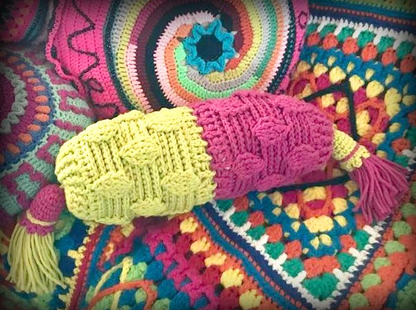 Gelb-rosa gehäkeltes Rollen-Kissen mit Quasten auf bunten Granny-Square-Decken