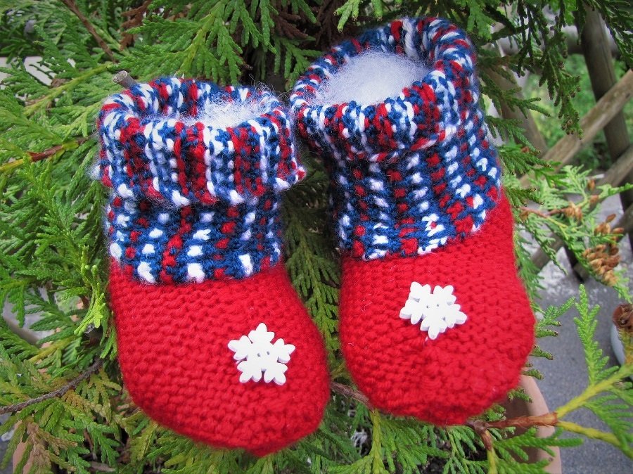 Zwei handgestrickte Babyschuhe in Rot, Blau und Weiß mit weißen Schneeflocken, liegend auf Nadelbaumzweigen.