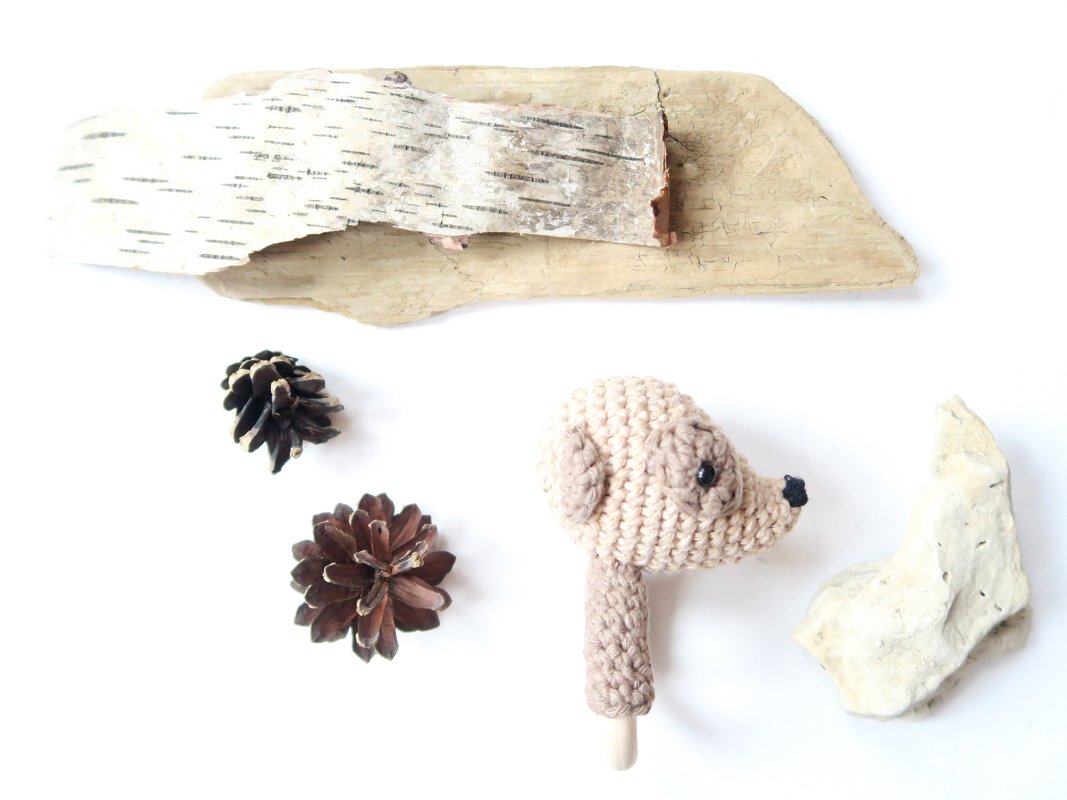 Gehäkelte Babyrassel in Tierkopfform mit Holzgriff, umgeben von Zapfen, Birkenrinde und Treibholz auf weißem Hintergrund.