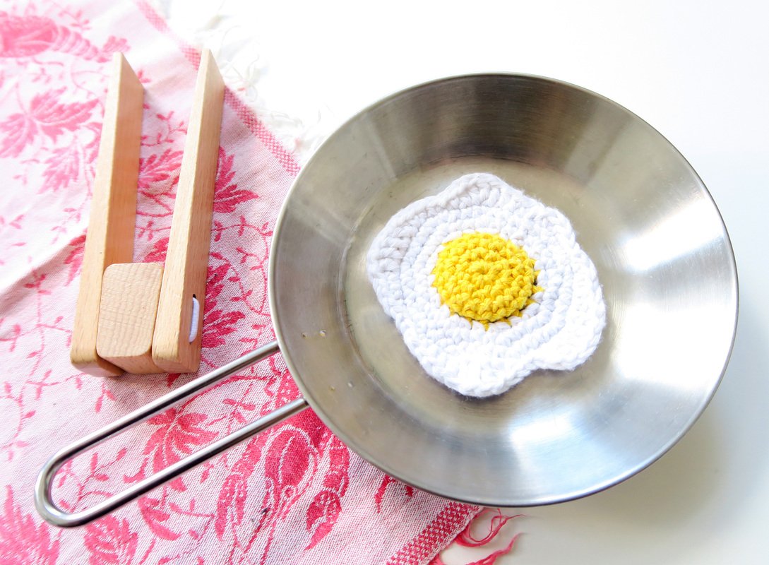 Gehäkeltes Spiegelei in Metallpfanne auf rosa Stoff neben Holzklammer