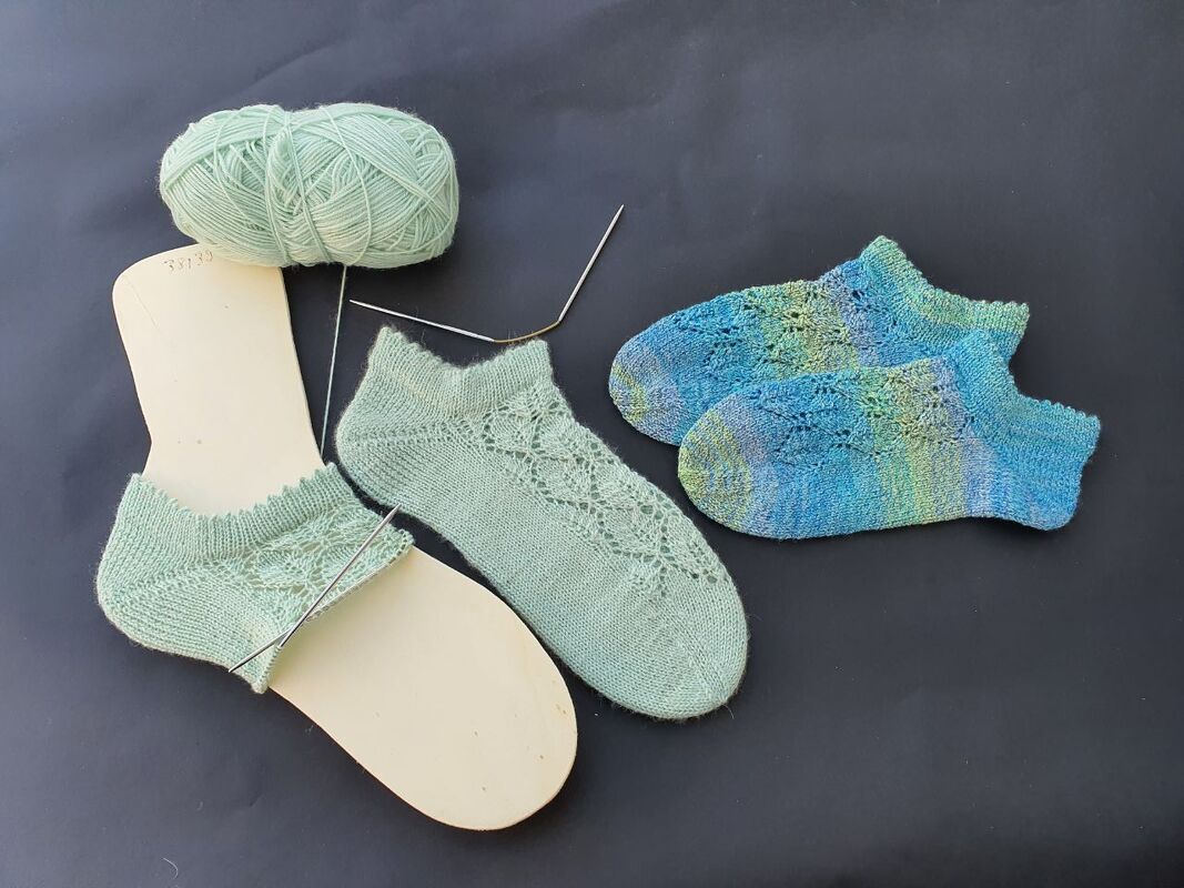 Zwei kurze gestrickte Füsslinge mit Lochmuster; mintgrünes Paar auf Sockenbrett mit Stricknadel und Wollknäuel, daneben ein mehrfarbiges Paar.