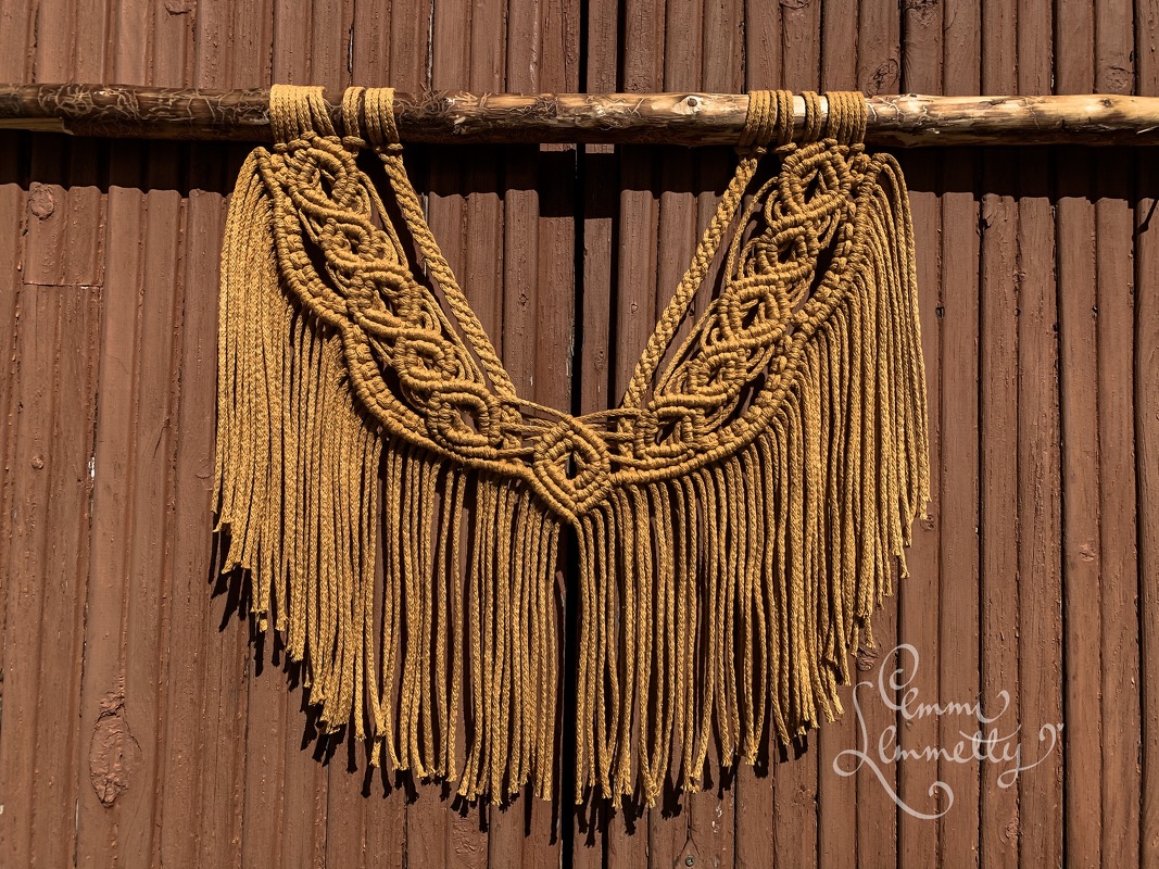 Mustard macramé wall hanging with braided knotwork and long fringe on a wooden dowel against a brown wooden wall