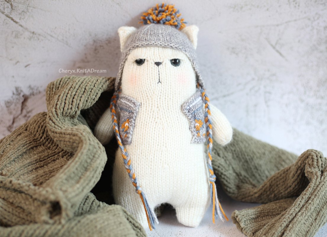 White knitted amigurumi cat wearing a grey winter hat with orange pompom, braided ties and a grey patterned vest on a green knit blanket
