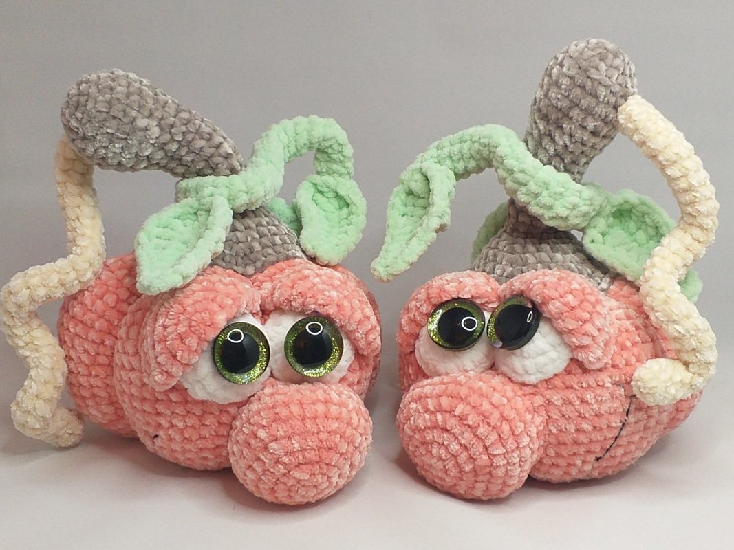 Two peach-colored crocheted amigurumi pumpkins with large green glittery eyes, layered leaf accents and pale grey stems on a neutral background.