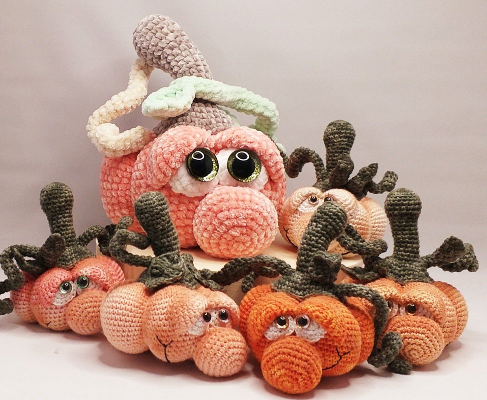 Group of amigurumi pumpkins: one large pastel pink and several smaller orange and peach with expressive eyes and green stems.