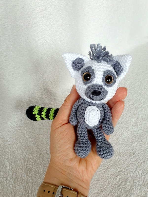 Hand hält kleines gehäkeltes Katta-Lemur-Amigurumi mit grau-weißem Gesicht und schwarz-grün gestreiftem Ringelschwanz.