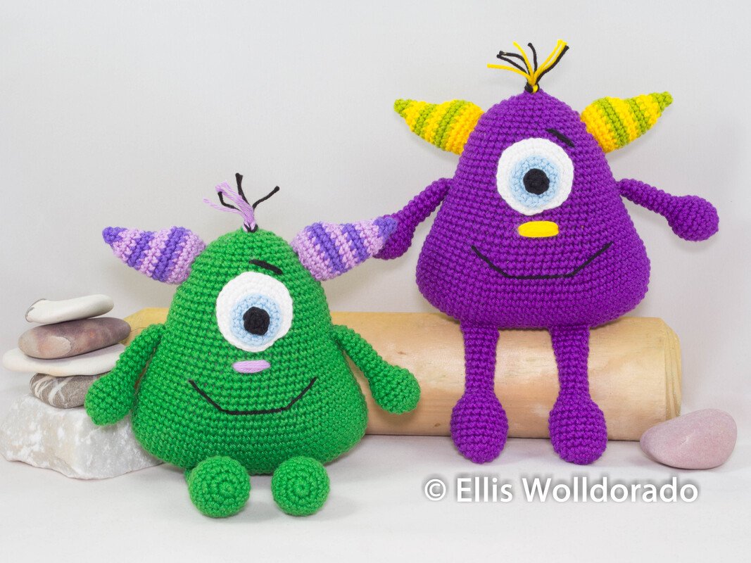 Zwei gehäkelte Amigurumi-Monster (grün und lila) mit je einem Auge und gestreiften Ohren auf Dekosteinen und Holz.