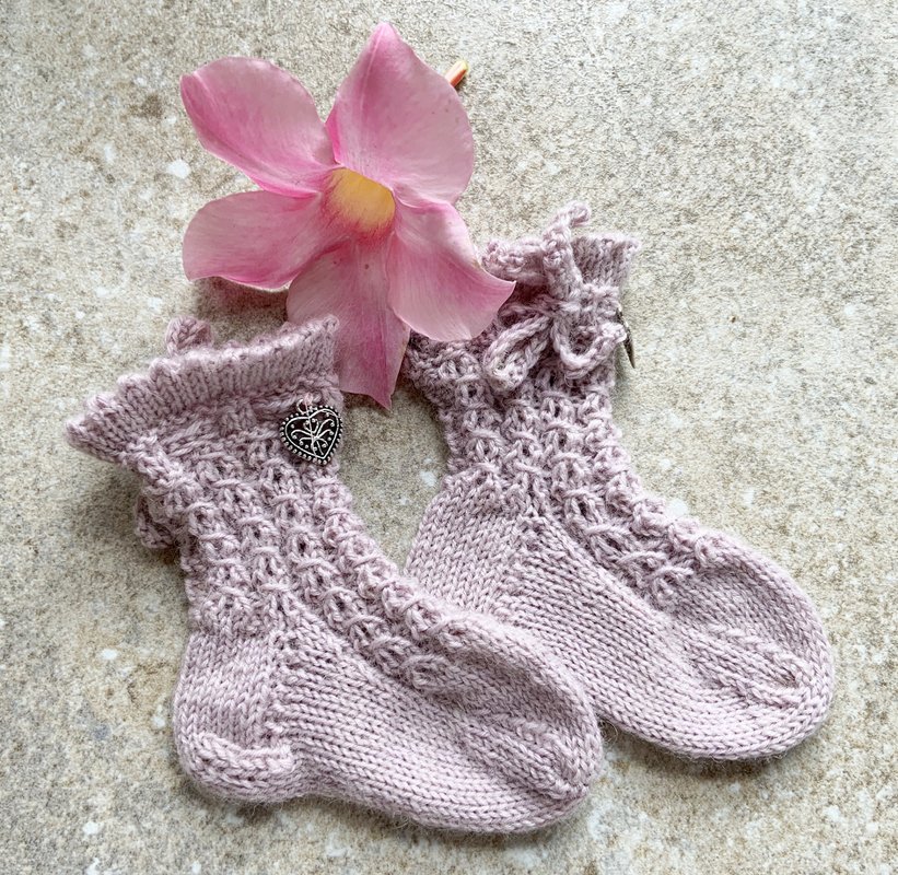 Zwei handgestrickte Babysocken in Lavendelfarben mit Rüschenbündchen und Herzanhänger neben einer rosa Blüte auf hellem Untergrund.