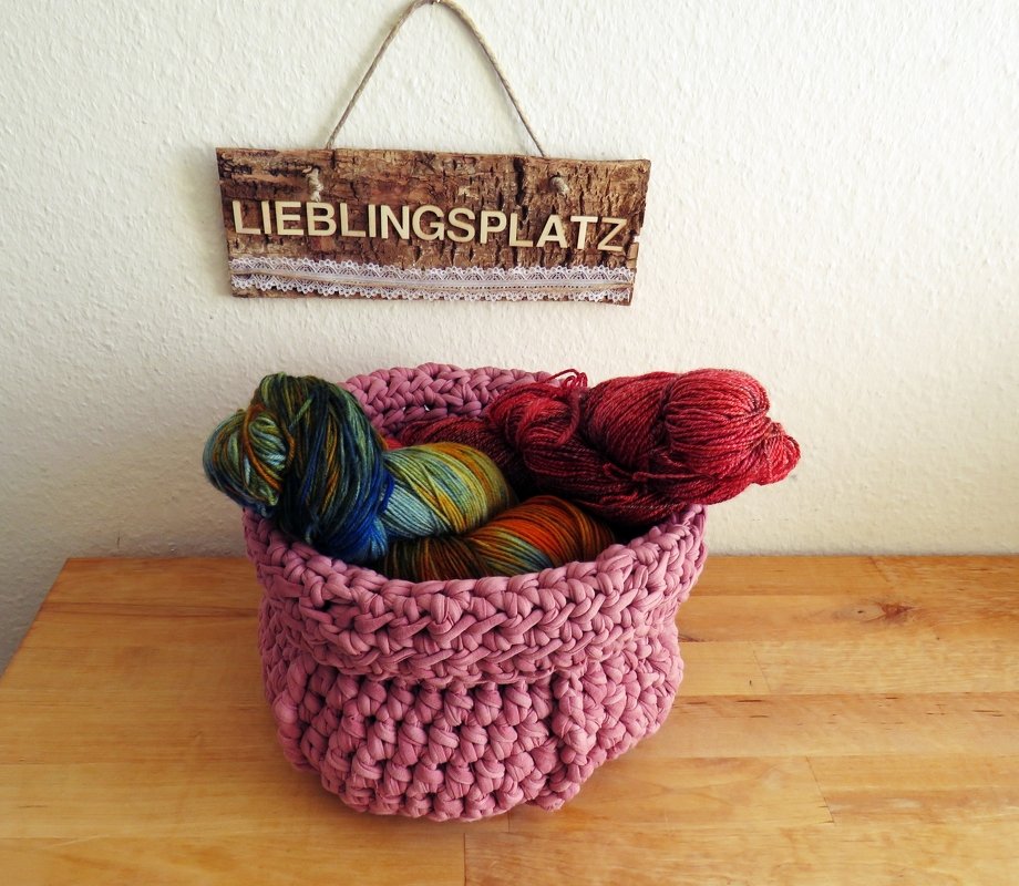 Rosa gehäkelter Korb aus Textilgarn, gefüllt mit bunten Wollknäueln, auf einem Holztisch; im Hintergrund Holzschild 'LIEBLINGSPLATZ'.