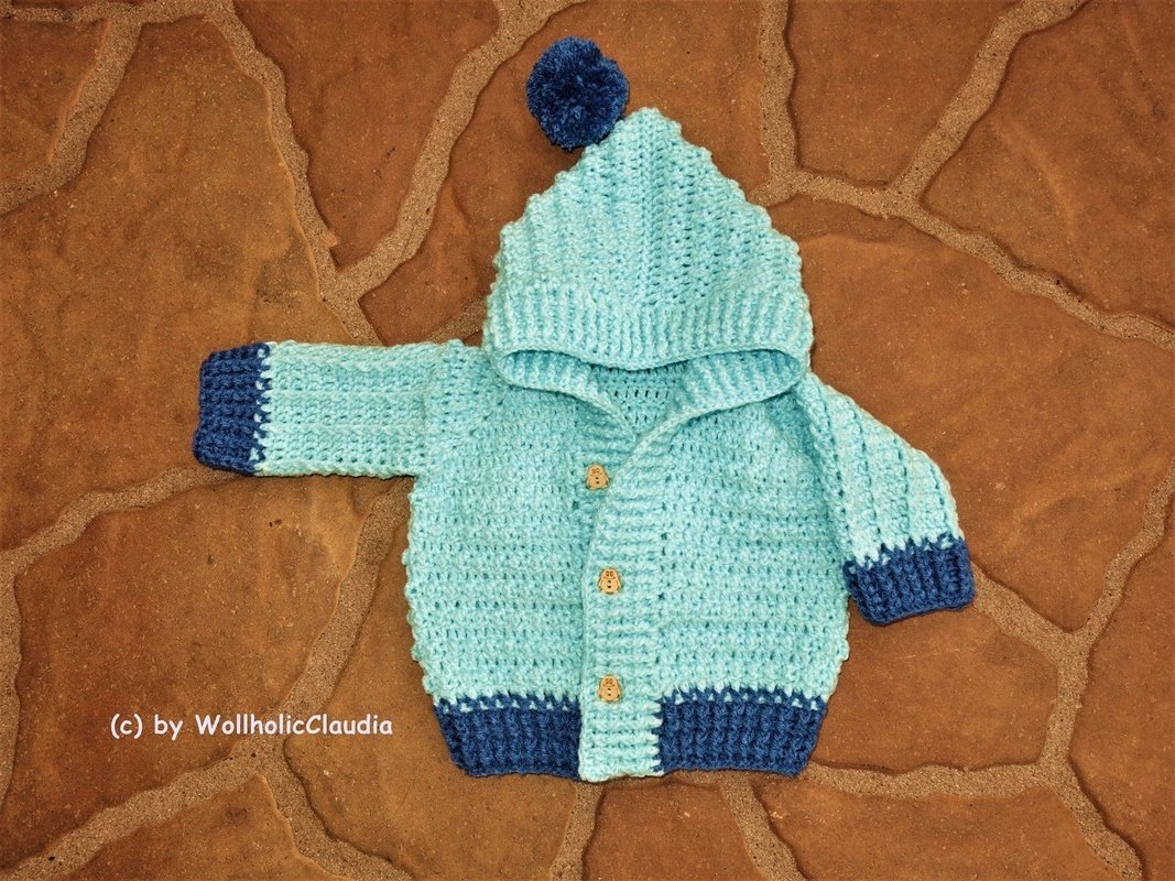 Hellblaue gehäkelte Babyjacke mit Kapuze, dunkelblaue Bündchen und Bommel auf steinfarbenem Untergrund