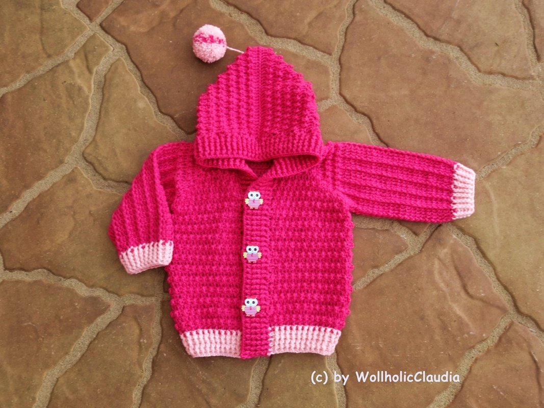 Fuchsiafarbene gehäkelte Babyjacke mit Kapuze und Eulenknöpfen auf braunem Steinboden