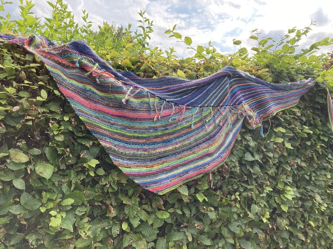 Buntes, gestreiftes Dreieckstuch mit Fransen an einer grünen Hecke