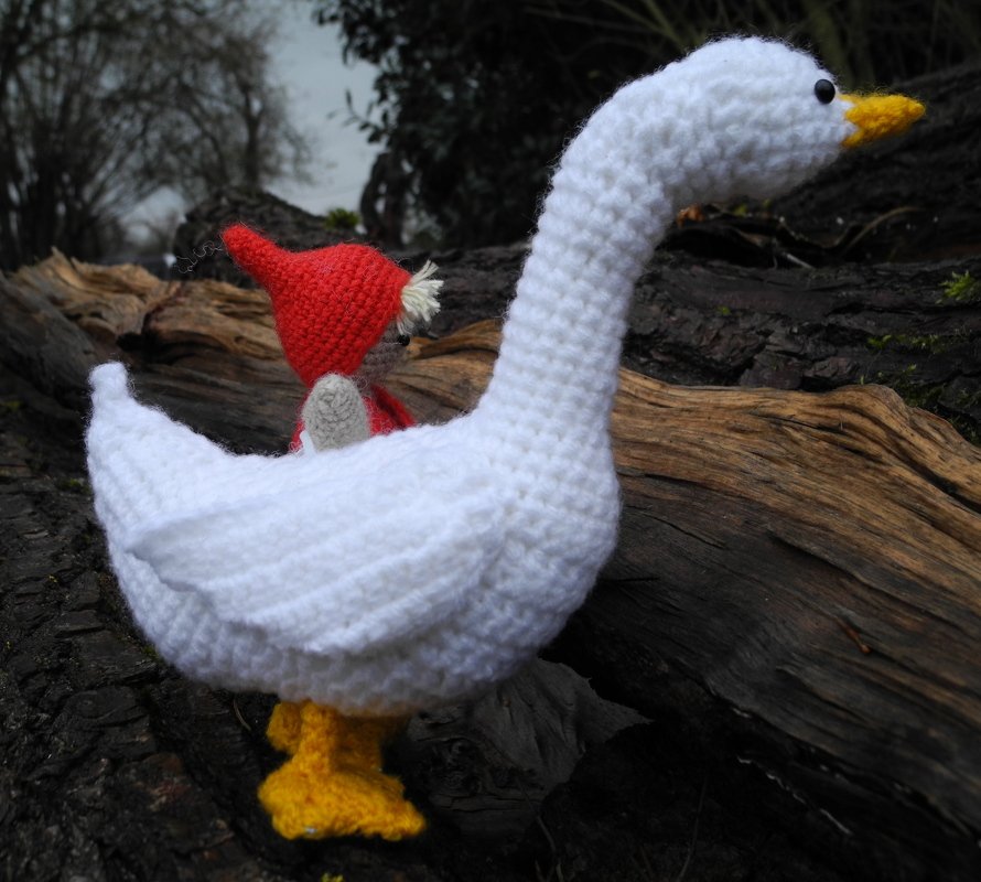 Weiß gehäkelte Gans mit gelbem Schnabel; kleine rote Zipfelmütze‑Figur sitzt auf dem Rücken