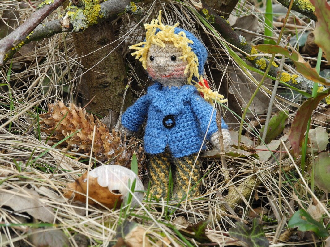 Gehäkelte Puppe mit blauer Jacke und gelbem Haar im Gras neben Zapfen