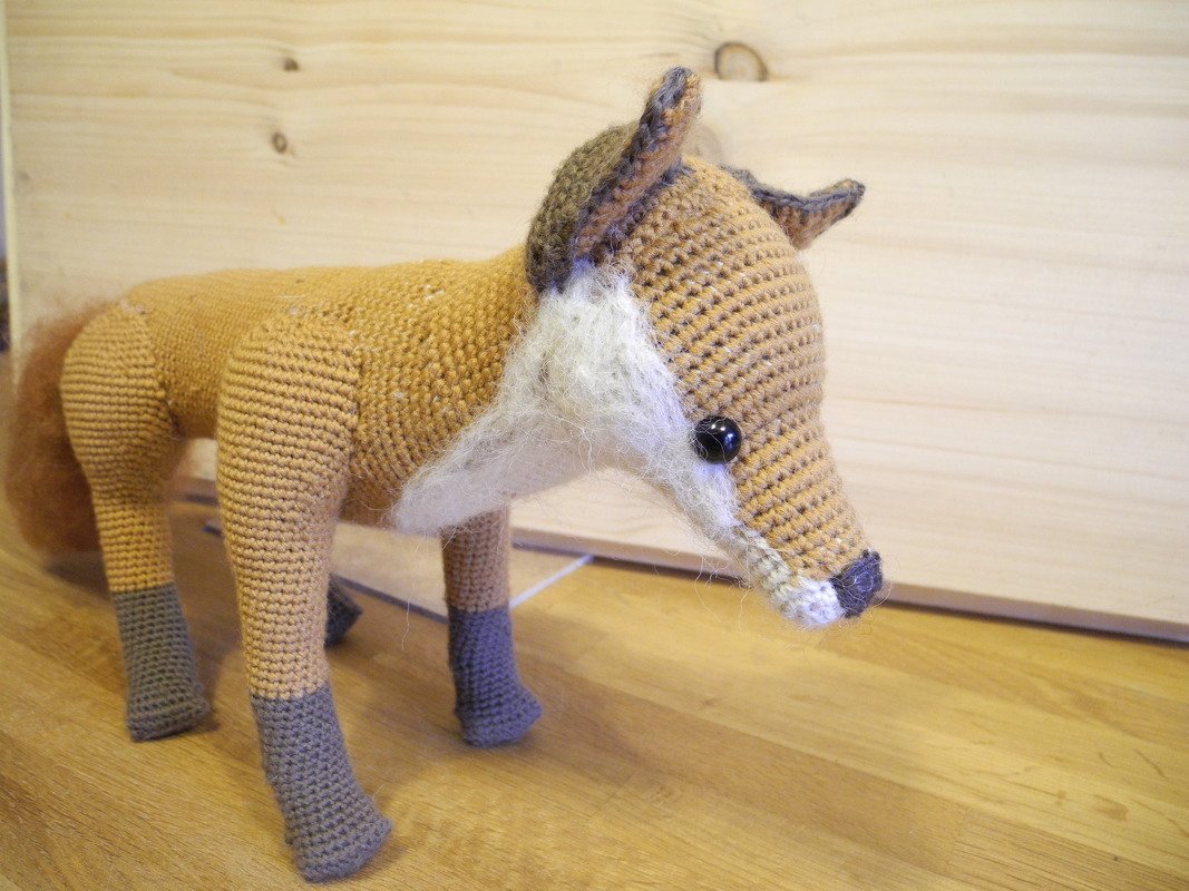 Gehäkelter Amigurumi-Fuchs in Braun- und Beigetönen mit weißer Schnauze und grauen Pfoten auf Holzuntergrund.
