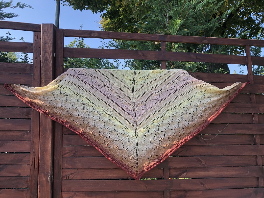 Gestricktes Dreieckstuch in Gelb-, Rosa- und Burgundertönen mit Lochmuster, über einem Holzzaun drapiert
