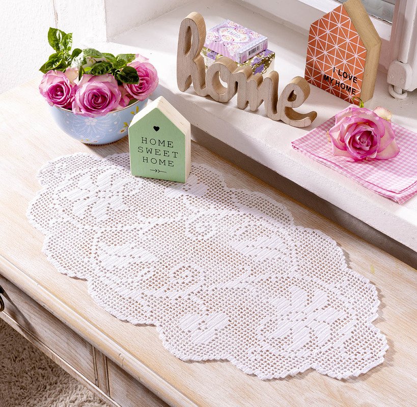 Weißer gehäkelter Tischläufer auf einer hellen Holzkommode, dekoriert mit rosa Rosen, einem Holzschriftzug und kleinen Dekoobjekten.