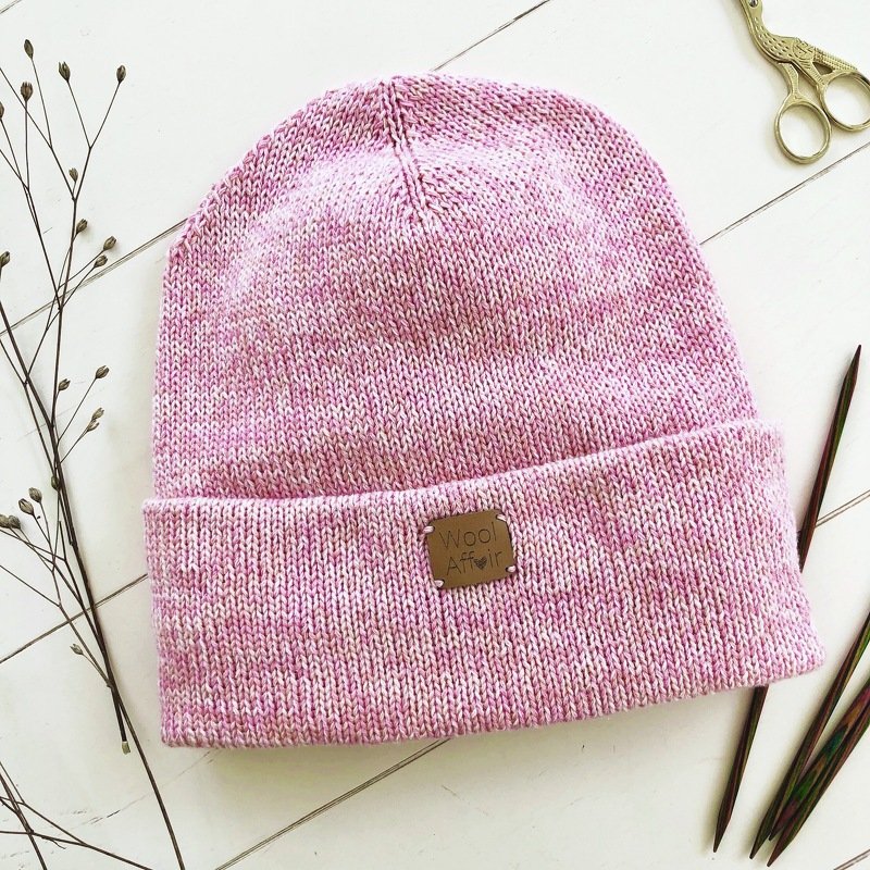 Pink speckled knitted beanie with a ribbed folded cuff laid flat on a white wooden surface with dried stems, knitting needles and scissors nearby.