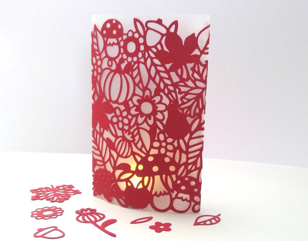 Rote, fein geschnittene Papierlaterne mit Pilz-, Blumen- und Blattmotiven, innen beleuchtet; ausgeschnittene Dekoteile liegen auf weißem Untergrund