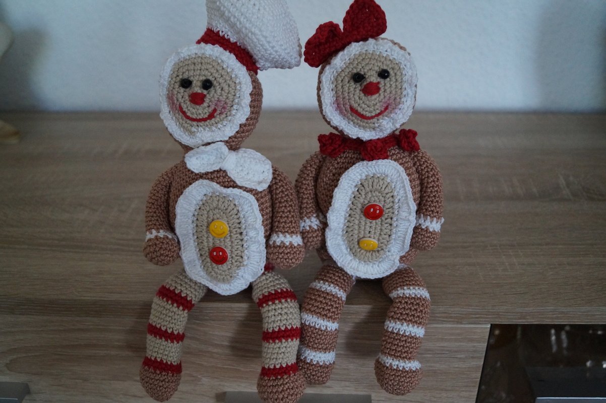 Zwei gehäkelte Lebkuchenfiguren auf einer Holzfläche, eine mit weißer Kopfbedeckung, die andere mit roter Schleife