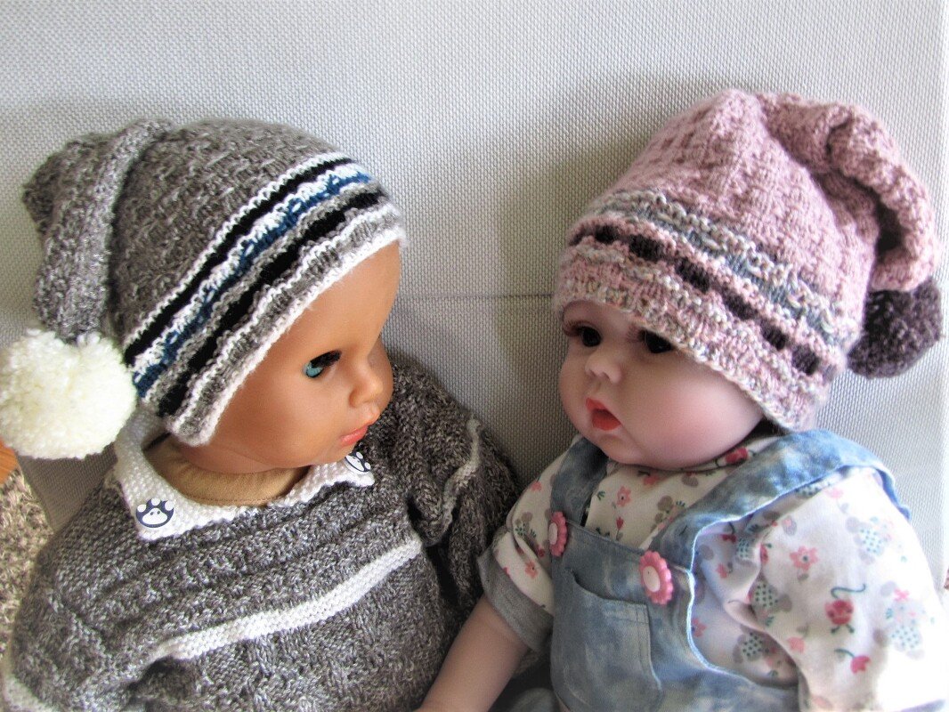Zwei Puppen mit gestrickten Mützen und Pullovern, links grau mit Bommel, rechts rosa gemustert