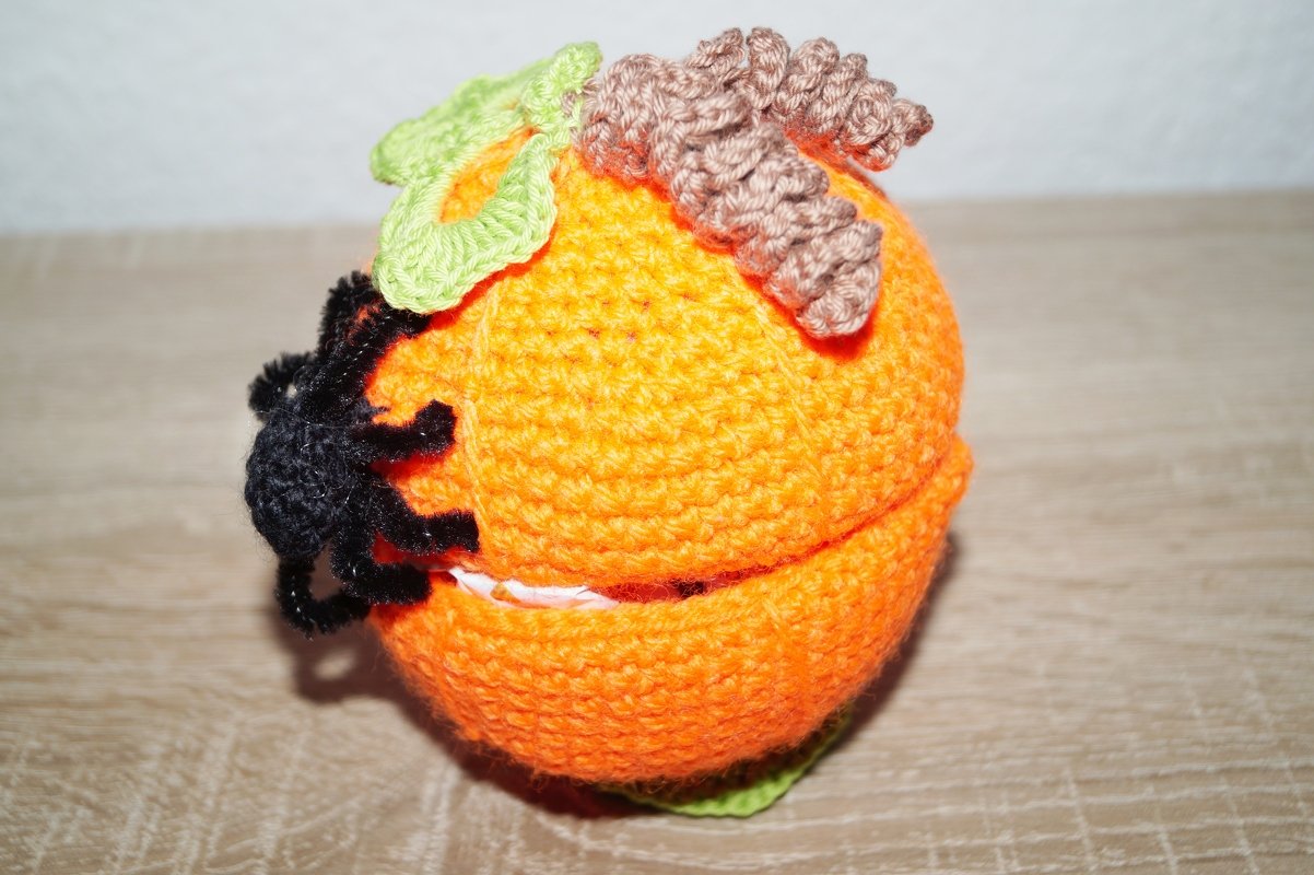 Gehäkelter orangefarbener Kürbis mit braunem Stiel, grünen Blättern und schwarzer Spinne; Utensilo leicht geöffnet.