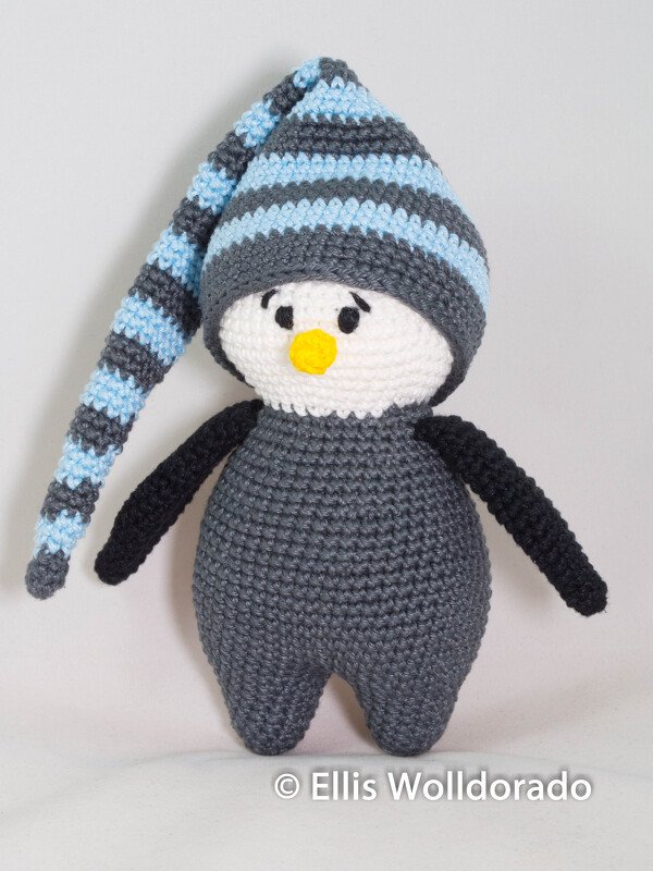 Gehäkelter Amigurumi-Pinguin, dunkelgrauer Körper, weißes Gesicht, gelber Schnabel und blau-grau gestreifte lange Mütze.