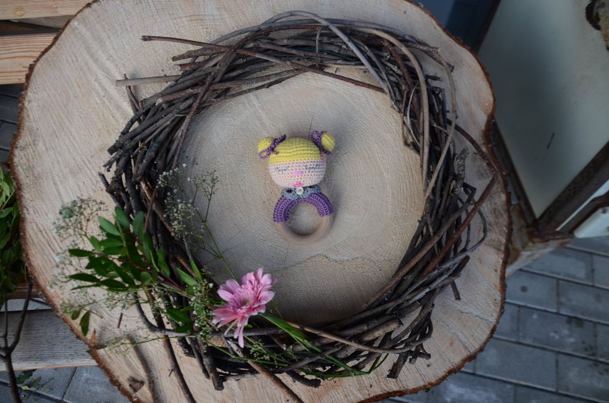 Gehäkelte Baby-Rassel mit gelben Zöpfchen und lila Oberteil auf Holzring, platziert auf einer Baumscheibe mit Zweigkranz und rosa Blüte