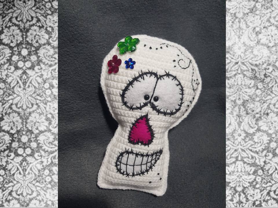 Weißer, gehäkelter Totenkopf mit schwarzen Sticknähten, rosa Filznase und bunten Blumenapplikationen vor dunklem Hintergrund.