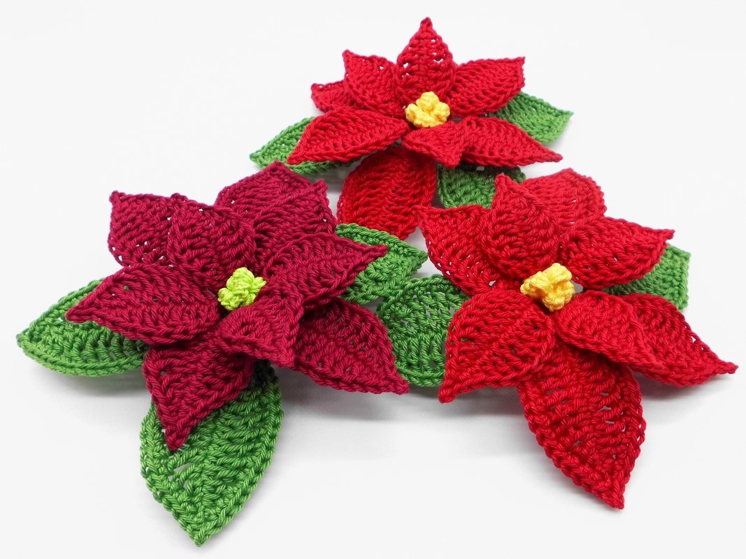 Three crocheted poinsettia motifs in red and burgundy with green leaves and yellow centers on a white background