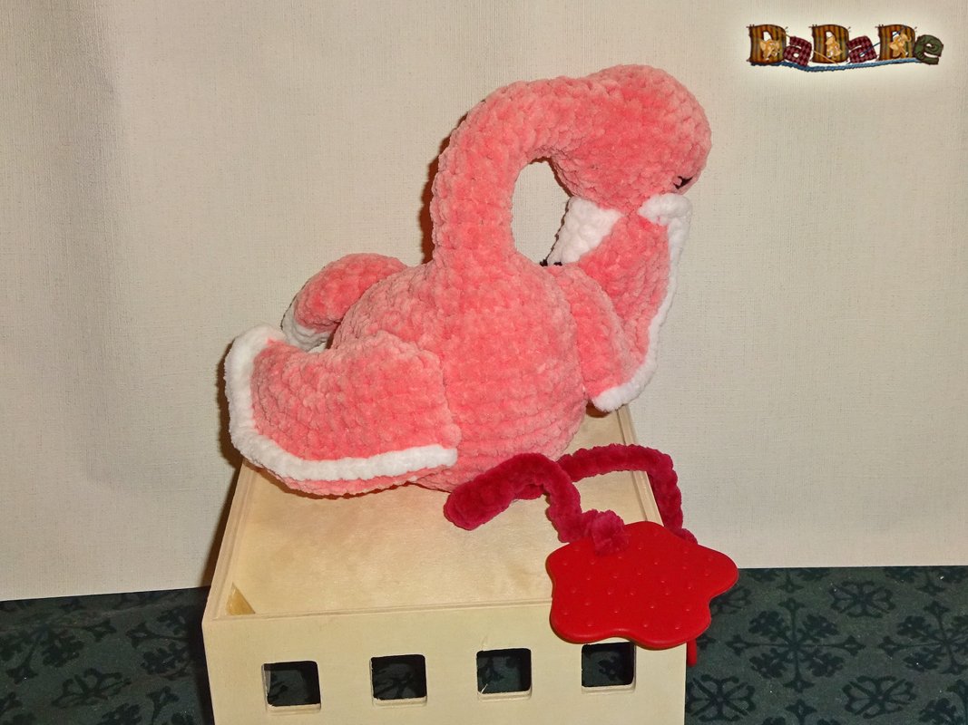 Rosa, handgehäkeltes Flamingo-Kuscheltier mit weißen Rändern an den Flügeln, auf einer hellen Holzbox, daneben ein rotes Beißspielzeug.