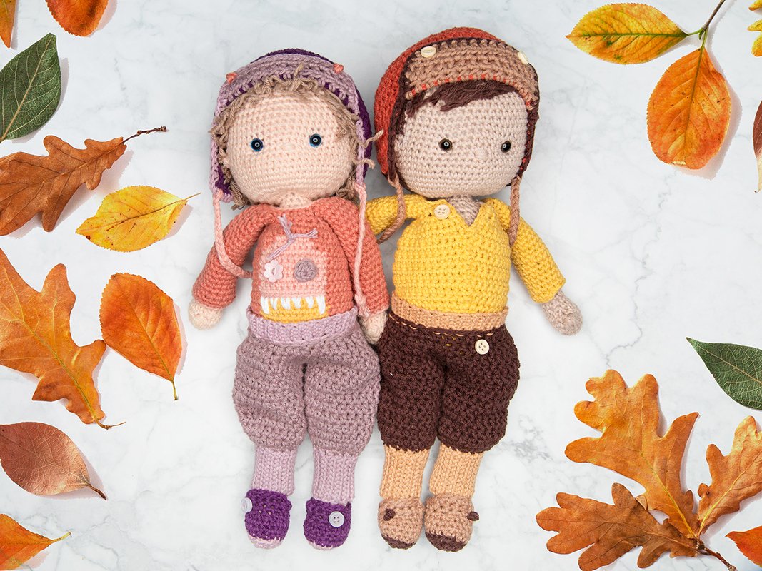 Zwei gehäkelte Amigurumi-Figuren mit Mützen und handgefertigter Kleidung, auf hellem Untergrund mit Herbstblättern gelegt.