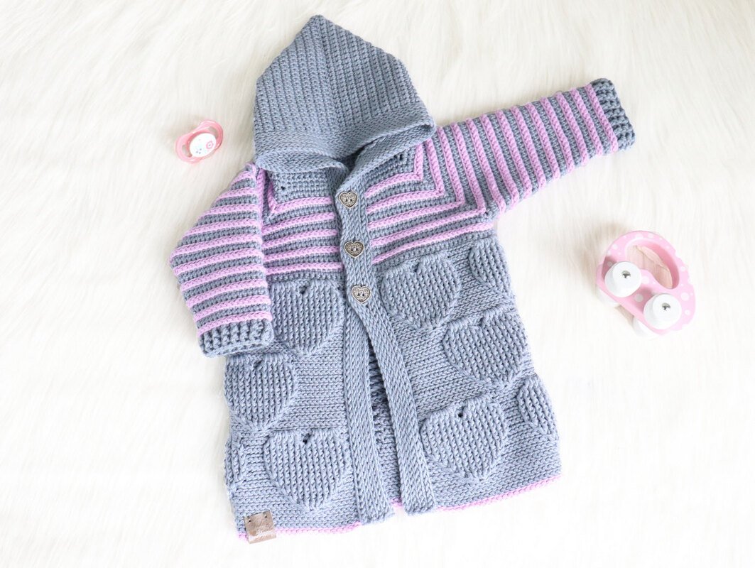 Grau-rosa gehäkelte Kapuzenjacke für Babys mit Streifen, Herztaschen und Herzknöpfen, liegend neben Schnuller und Spielzeug.