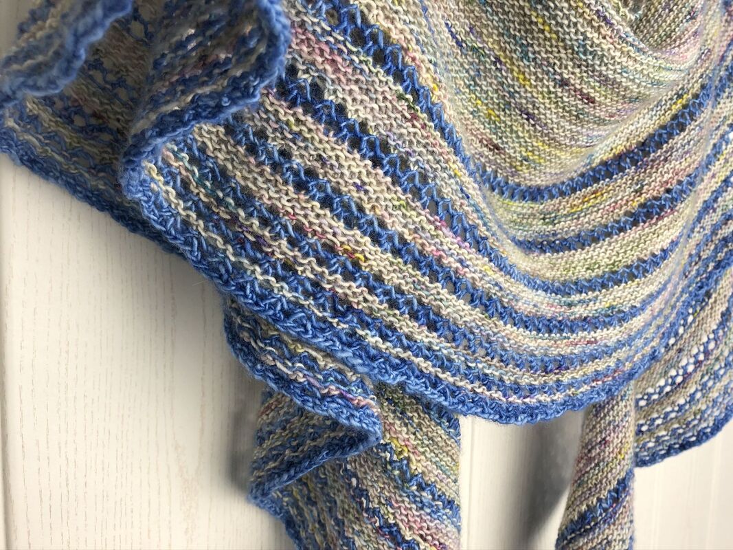 Close-up of a crescent garter-stitch shawl in speckled neutral yarn with blue stripes and scalloped blue edge