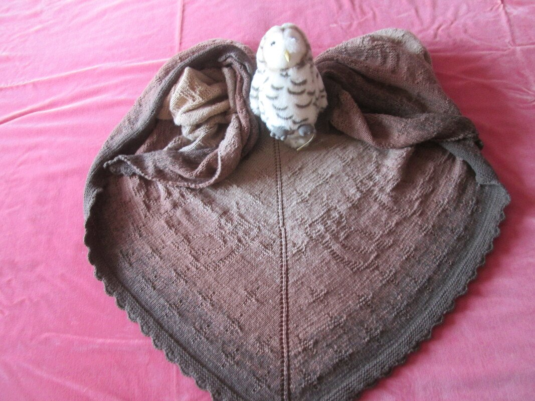 Braunes gestricktes Dreieckstuch mit Mittelnaht, Loch- und Reliefmuster auf rosafarbenem Untergrund, kleines Plüschtier an der Spitze