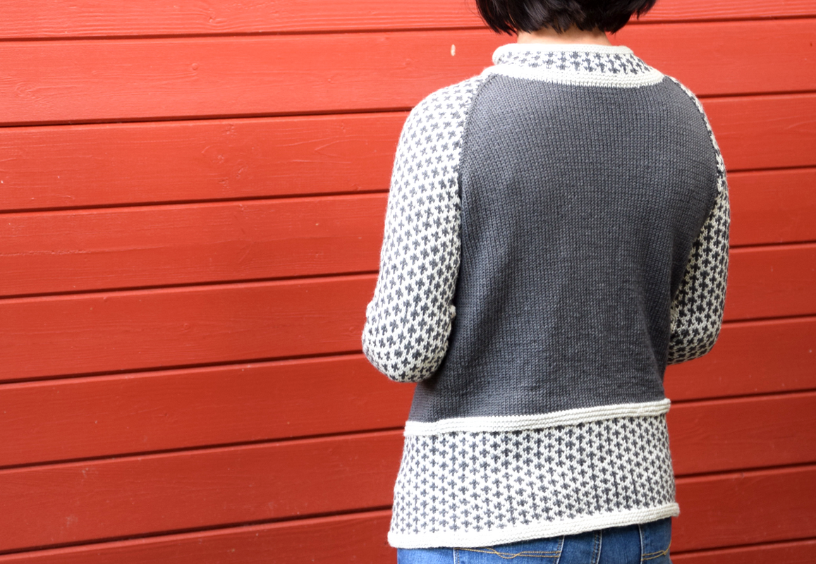 Rückenansicht eines grau-weißen Strickpullovers mit Jacquardärmeln vor roter Holzwand