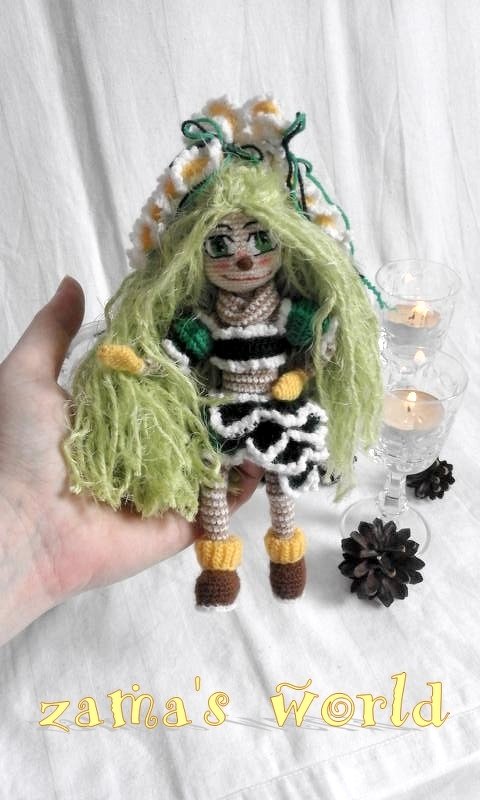 Hand hält gehäkelte Amigurumi-Puppe mit langem grünem Haar und weißen Blüten, schwarz-weißem Kleid und gelben Details