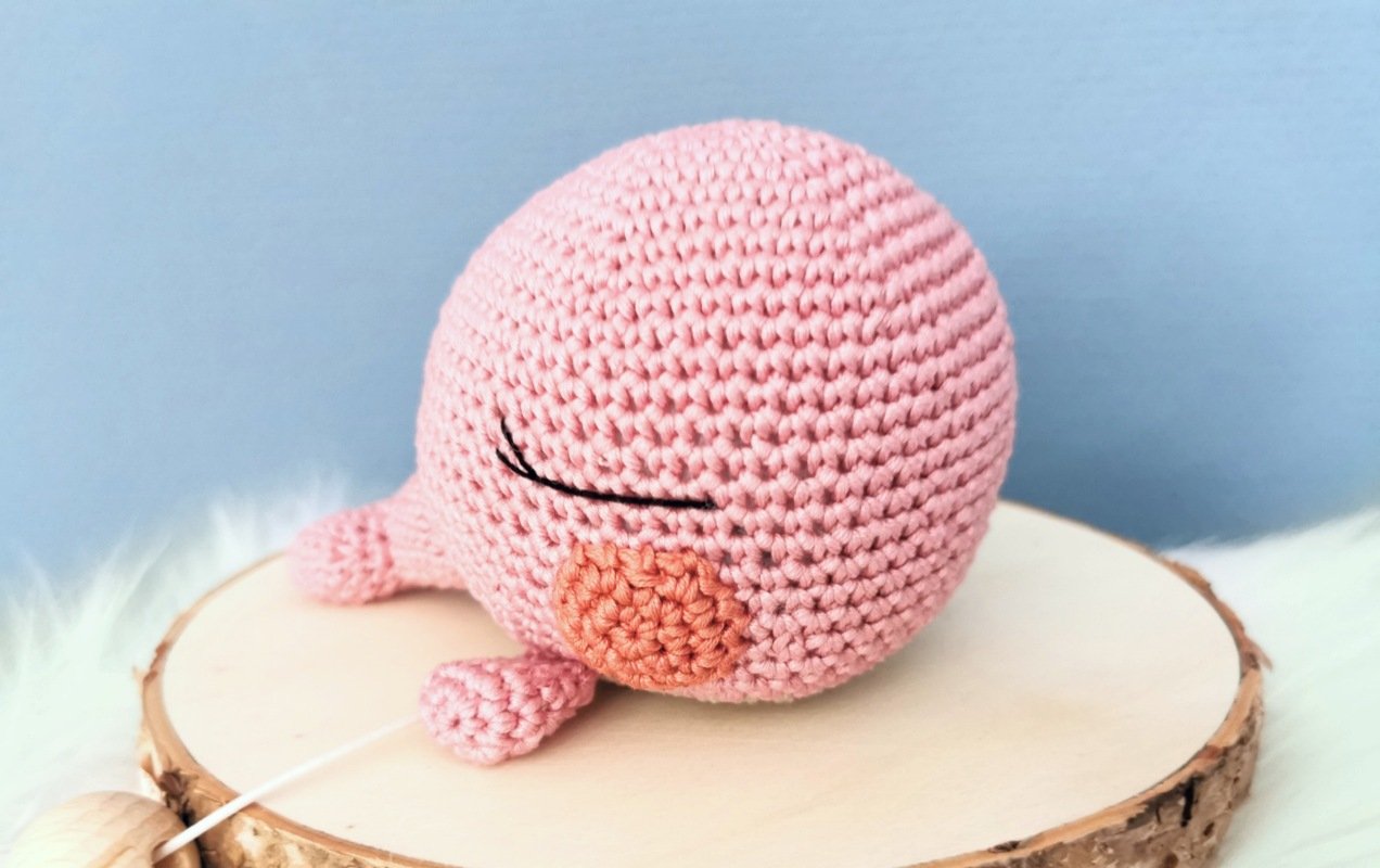 Rosa gehäkeltes Amigurumi mit geschlossenen Augen, rosafarbener Wange und Holzperlen-Zugband